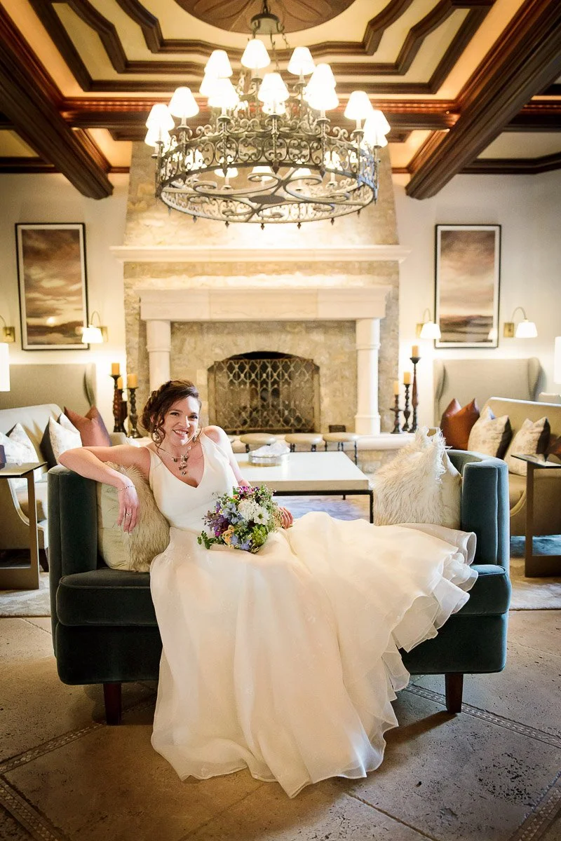 A smiling bride in a white gown lounges on a plush sofa, holding a colorful bouquet. The elegant room features a stone fireplace and ornate chandelier.