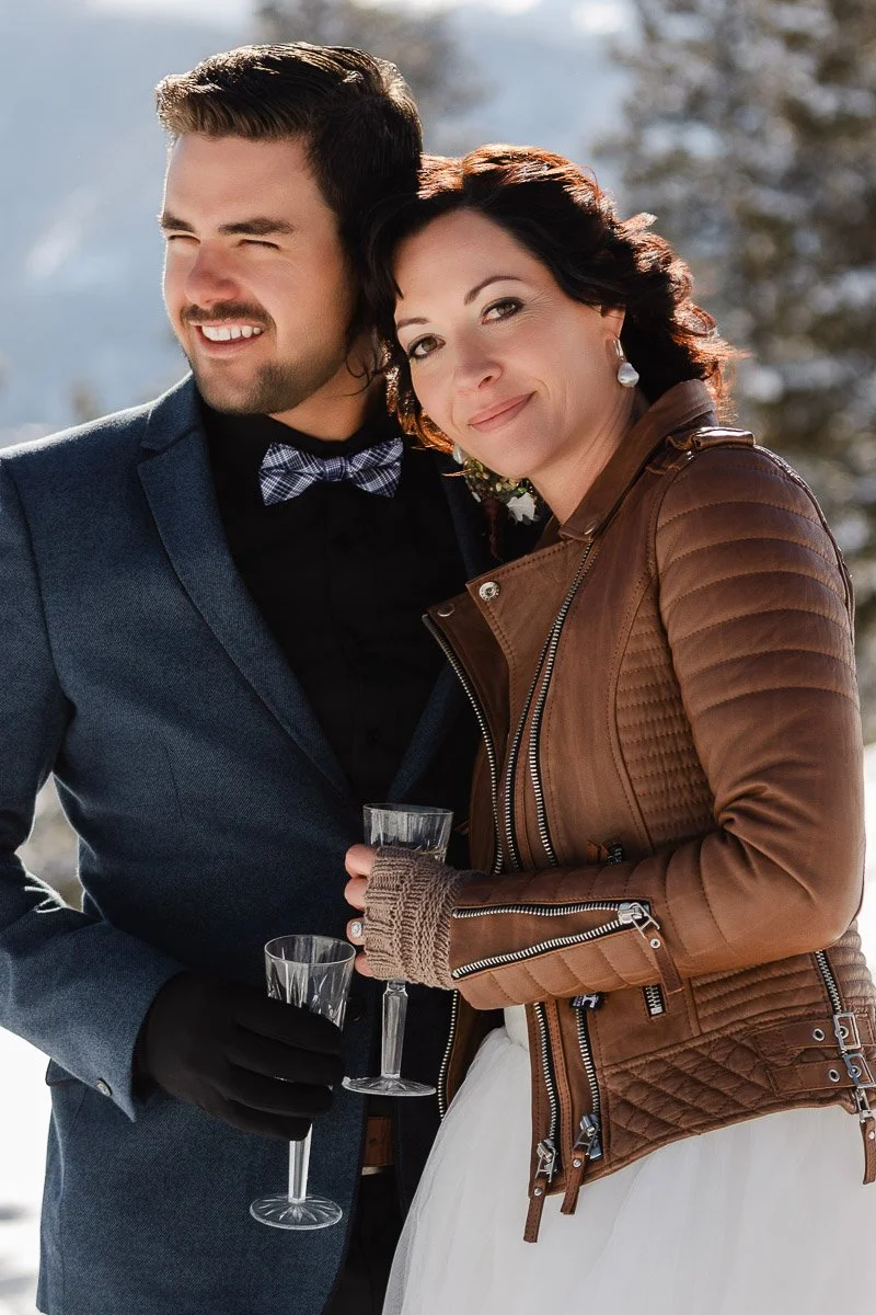 Smiling couple in winter outdoors; man in blue suit, woman in brown leather jacket over white dress, holding champagne glasses. Snowy background.