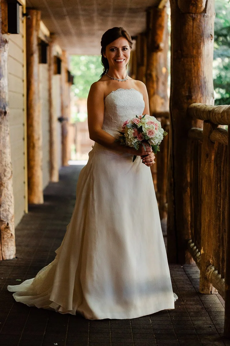 A bride in a strapless white gown stands smiling in a rustic hallway with wooden beams, holding a bouquet of pink and white flowers, conveying joy.