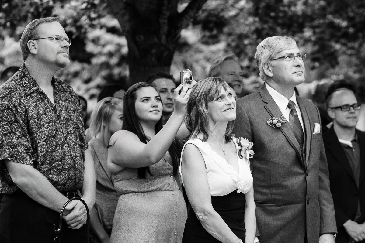A group of people stands outdoors, attentively watching something. A woman holds up a camera. The scene conveys anticipation and curiosity.