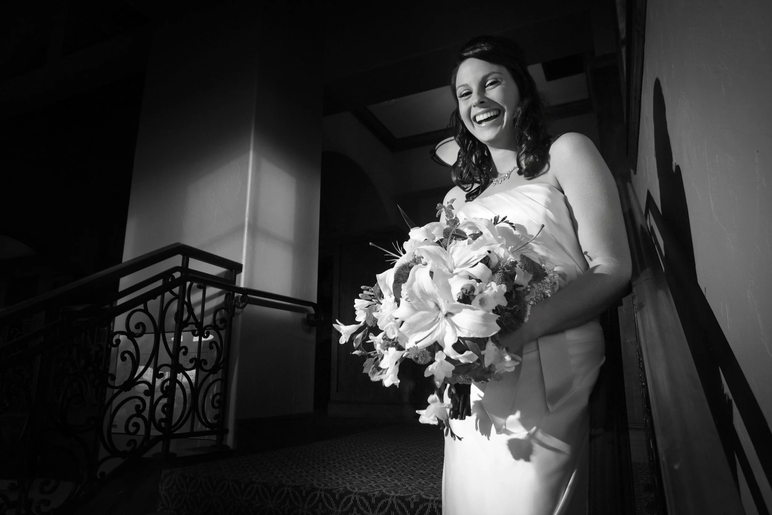 A joyful bride in a white gown holds a large bouquet of flowers, standing on a stairway. Dramatic black and white lighting enhances her laughter during a Valley Country Club wedding in Aurora, Colorado