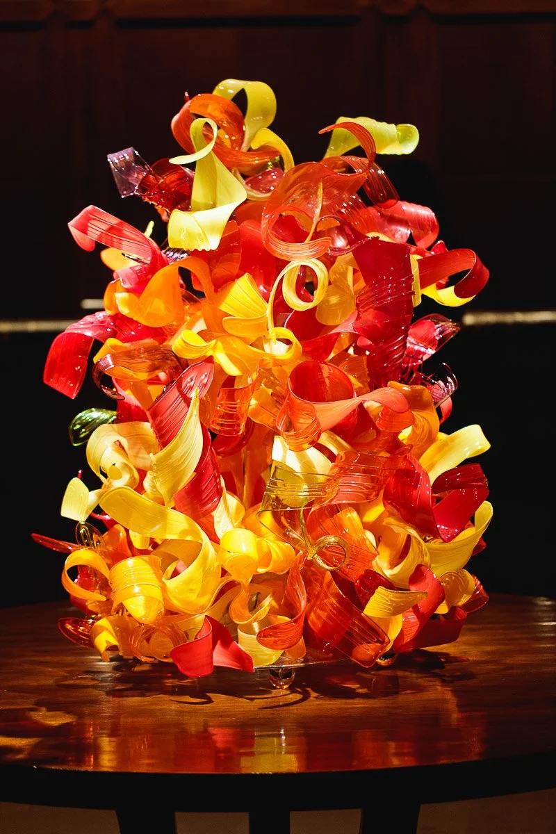 Vibrant glass sculpture with swirling red, orange, and yellow curls, sits atop a wooden table. The dynamic design exudes energy and creativity.