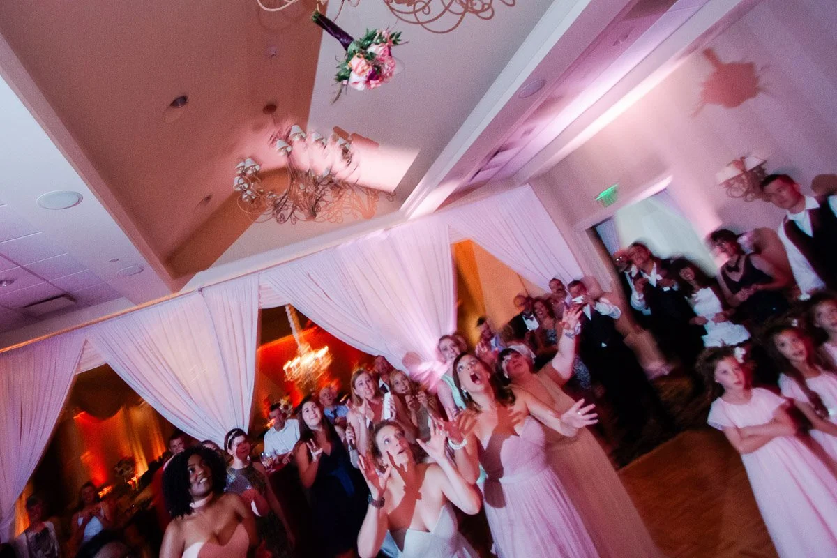 A bridal bouquet soars through the air, surrounded by excited women reaching up to catch it. They're in a decorated reception hall with pink lighting.
