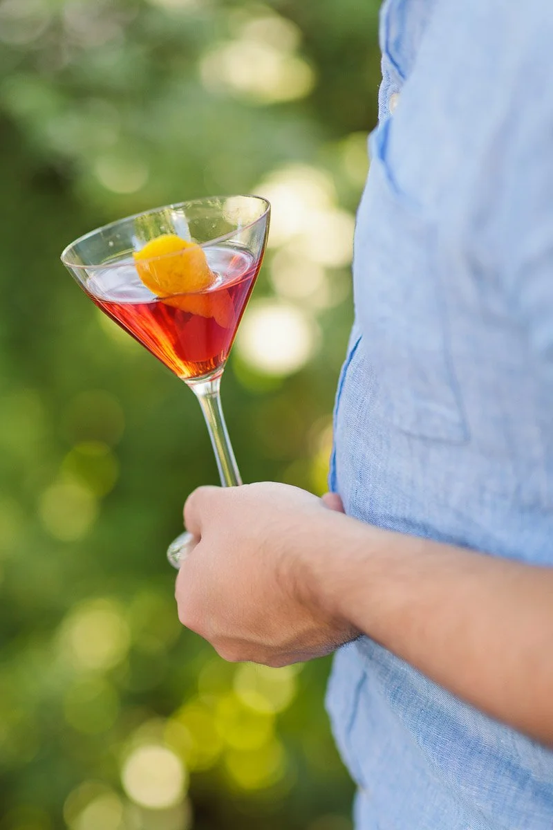 A person in a light blue shirt holds a martini glass with a red cocktail and orange garnish. The background is a lush, blurred greenery, creating a relaxed vibe.