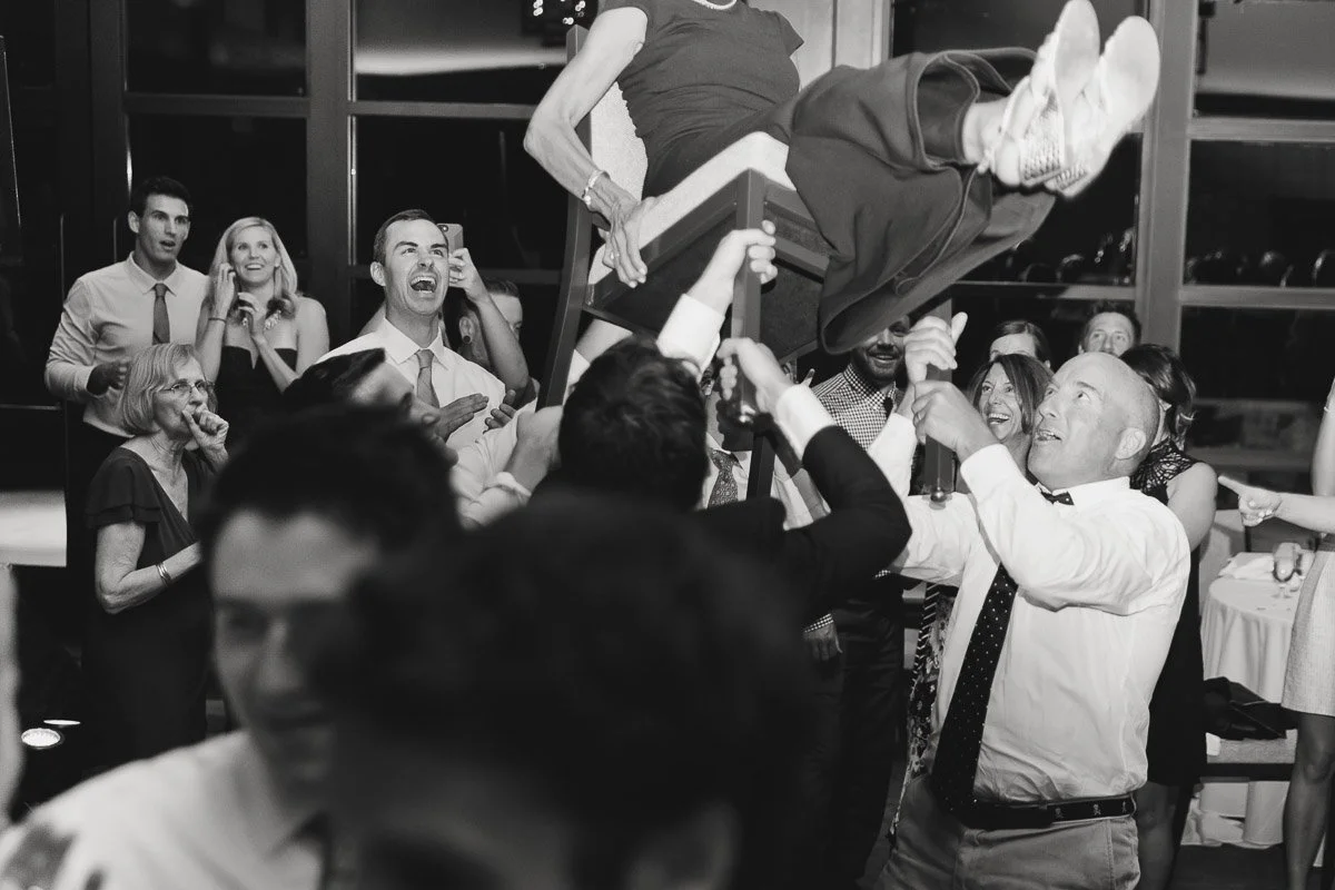 A black-and-white photo shows a lively party scene where a woman is being lifted on a chair by a group of smiling people, conveying joy and celebration.