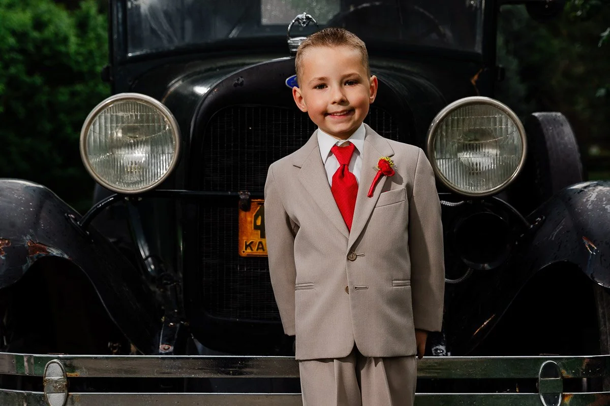 A young boy in a beige suit and red tie smiling confidently in front of a vintage 1929 Ford car. The background is a lush green setting, conveying a cheerful and nostalgic mood, captured by Chatfield Farms wedding photographer tomKphoto.