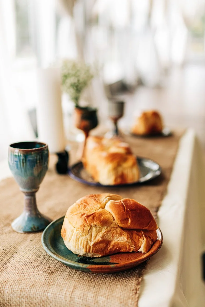 A rustic table setting features fresh bread on colorful ceramic plates, ornate goblets, and candles, creating an inviting and warm atmosphere.