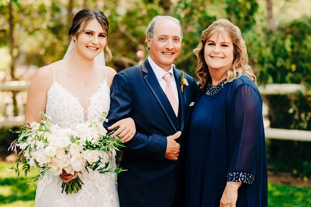 A bride in a white lace dress holds a bouquet, standing beside two older adults in formal attire, smiling in a garden setting.