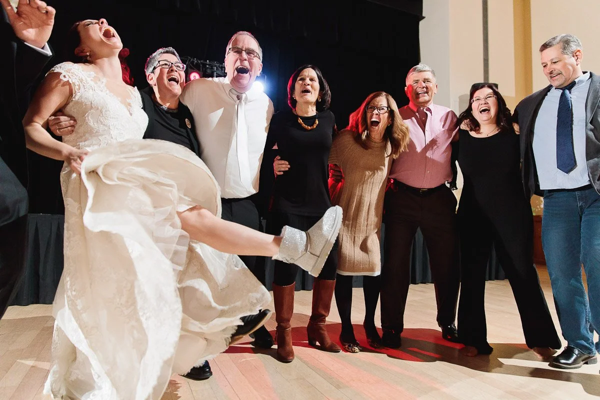 A joyful bride in a white dress kicks her leg out while dancing with seven people in semi-formal attire onstage, all smiling and laughing.
