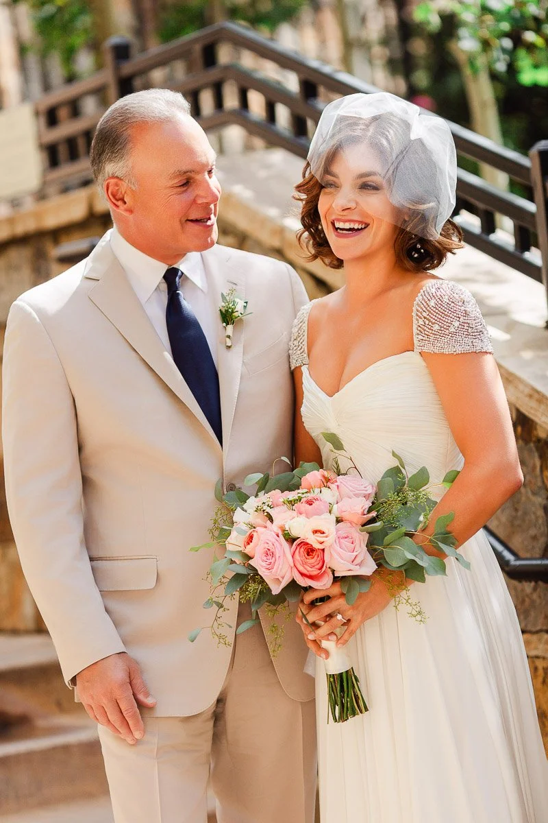 A smiling bride in an elegant white dress holds a pink rose bouquet, standing beside a man in a beige suit and blue tie. They appear joyful.