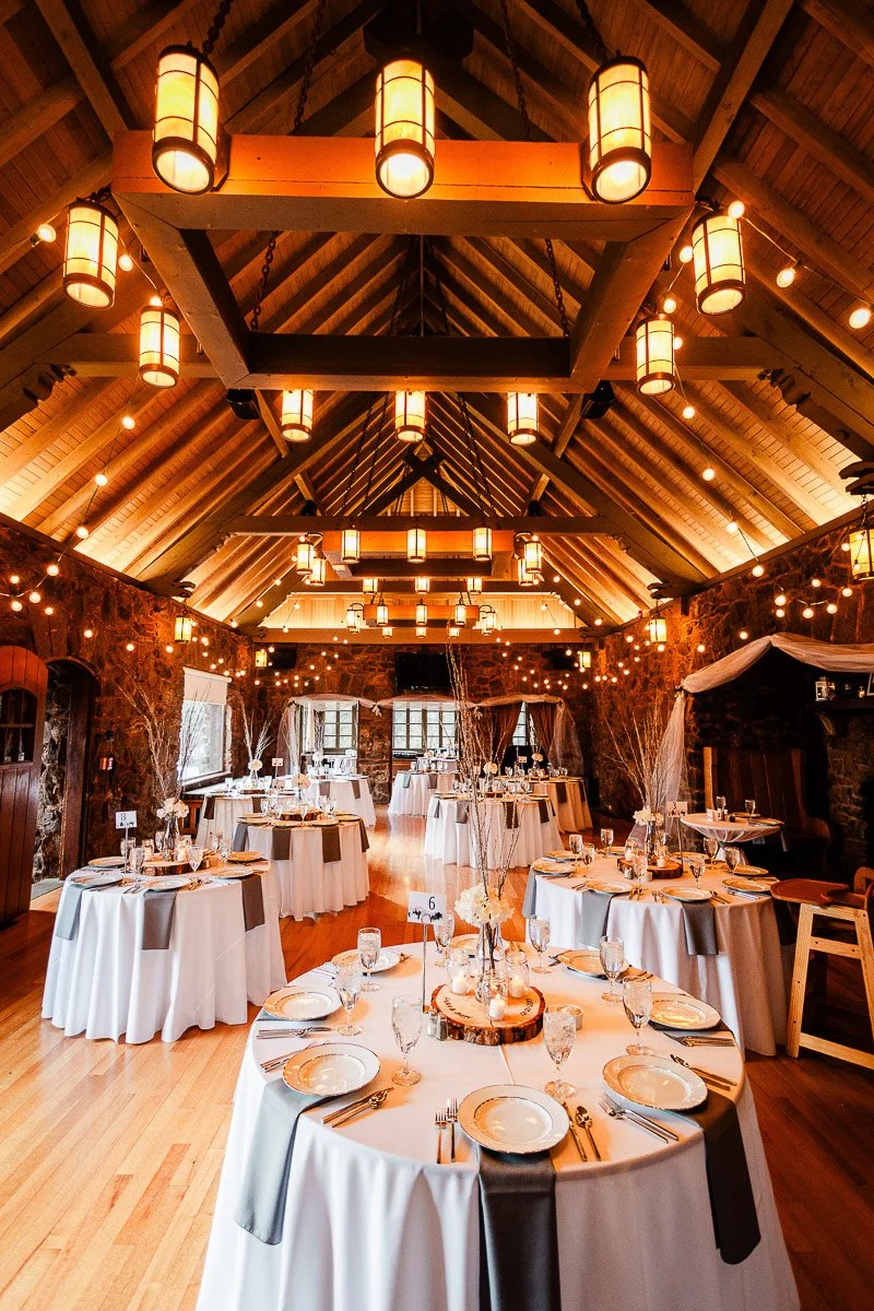 Elegant banquet hall with round tables set in white linens and gray napkins. Warm lighting from chandeliers and string lights creates a cozy ambiance captured by Boettcher Mansion wedding photographer tomKphoto