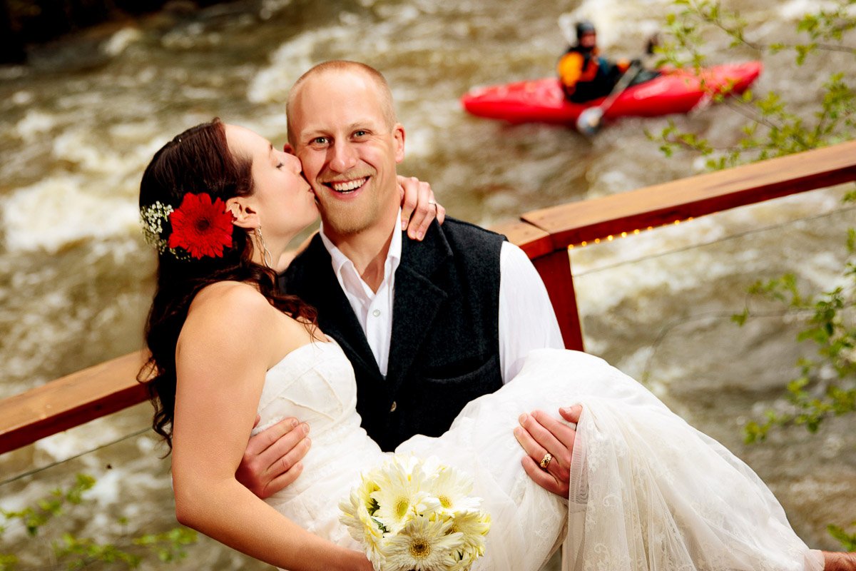 A bride in a white dress kisses a smiling groom holding her, with a vibrant red flower in her hair. A red kayak is seen on Boulder Creek in the background. Electric, joyous tone.