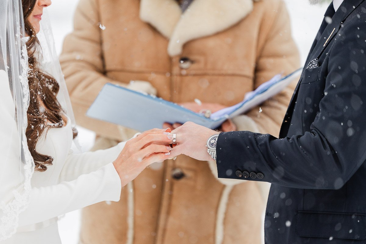 Bride and groom exchange rings in a snowy outdoor wedding ceremony. The bride wears a white dress, the groom a dark suit, while snowflakes fall gently.