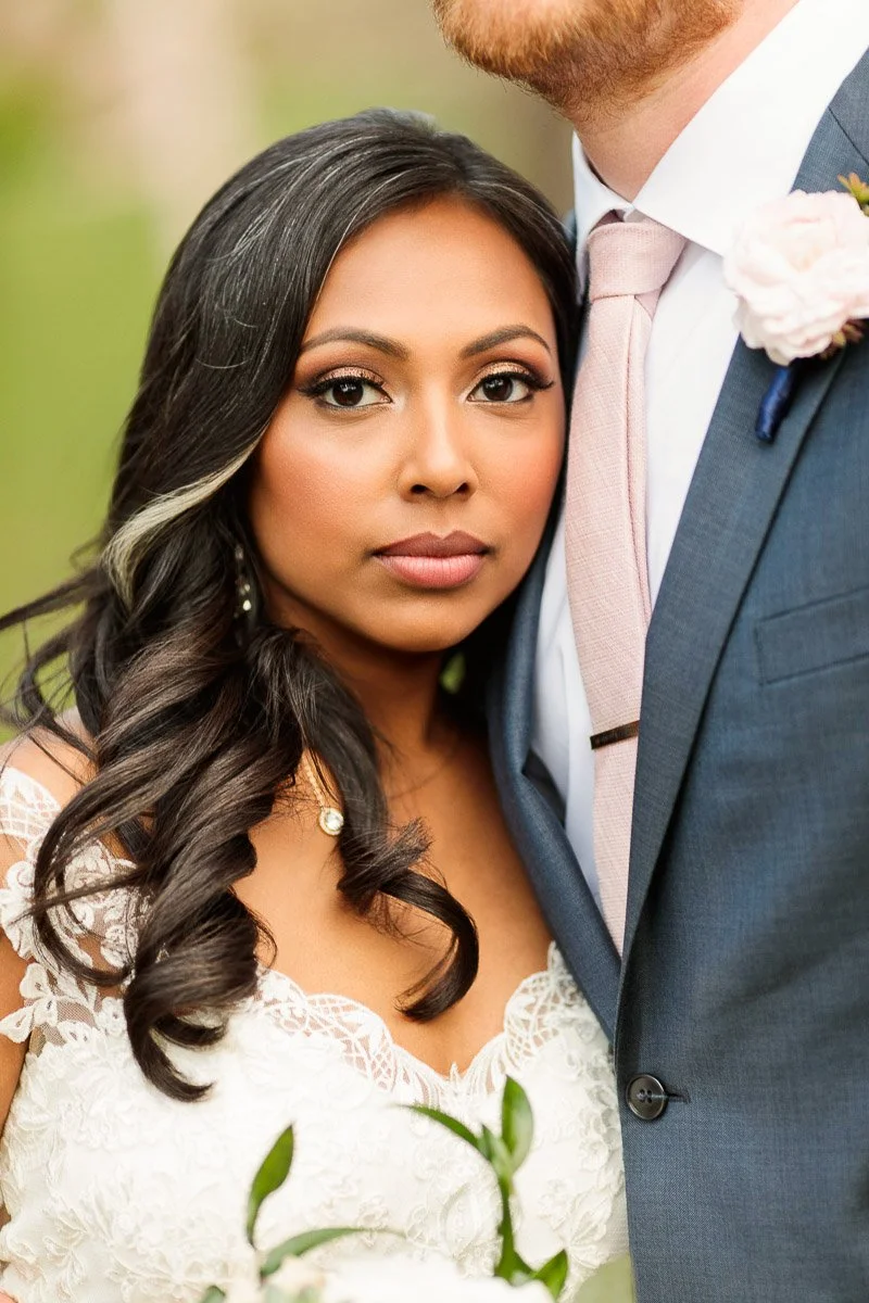 A bride in a lace dress stands beside a groom in a blue suit and pink tie, holding a bouquet. The scene conveys love and elegance.