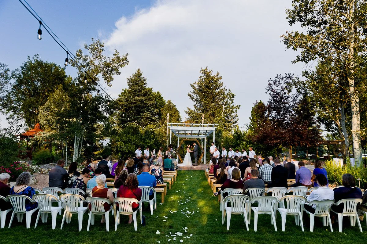 Outdoor Church Ranch Event Center wedding ceremony set in a garden, with a couple under an arbor, surrounded by guests seated on chairs. Trees and clear sky in the background.