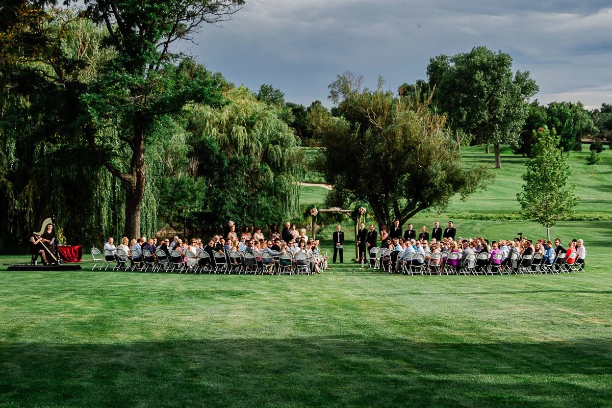 Outdoor Lakewood Country Club wedding ceremony on a lush green lawn, surrounded by trees. Guests sit in a semicircle, facing a couple under a floral arch. Cloudy sky overhead.
