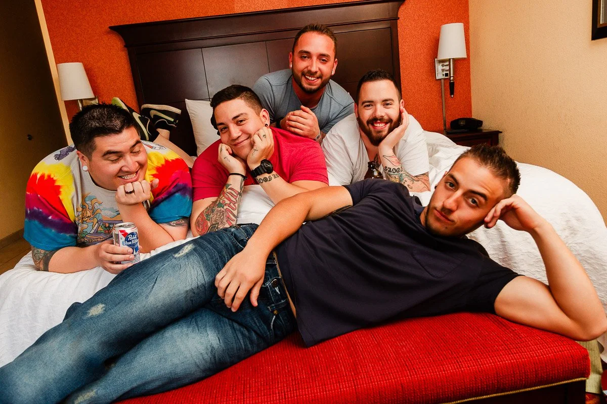 Five friends in a cozy hotel room pose playfully on a bed. Four are sitting, smiling happily, while one lounges in the foreground, creating a relaxed, joyful atmosphere.