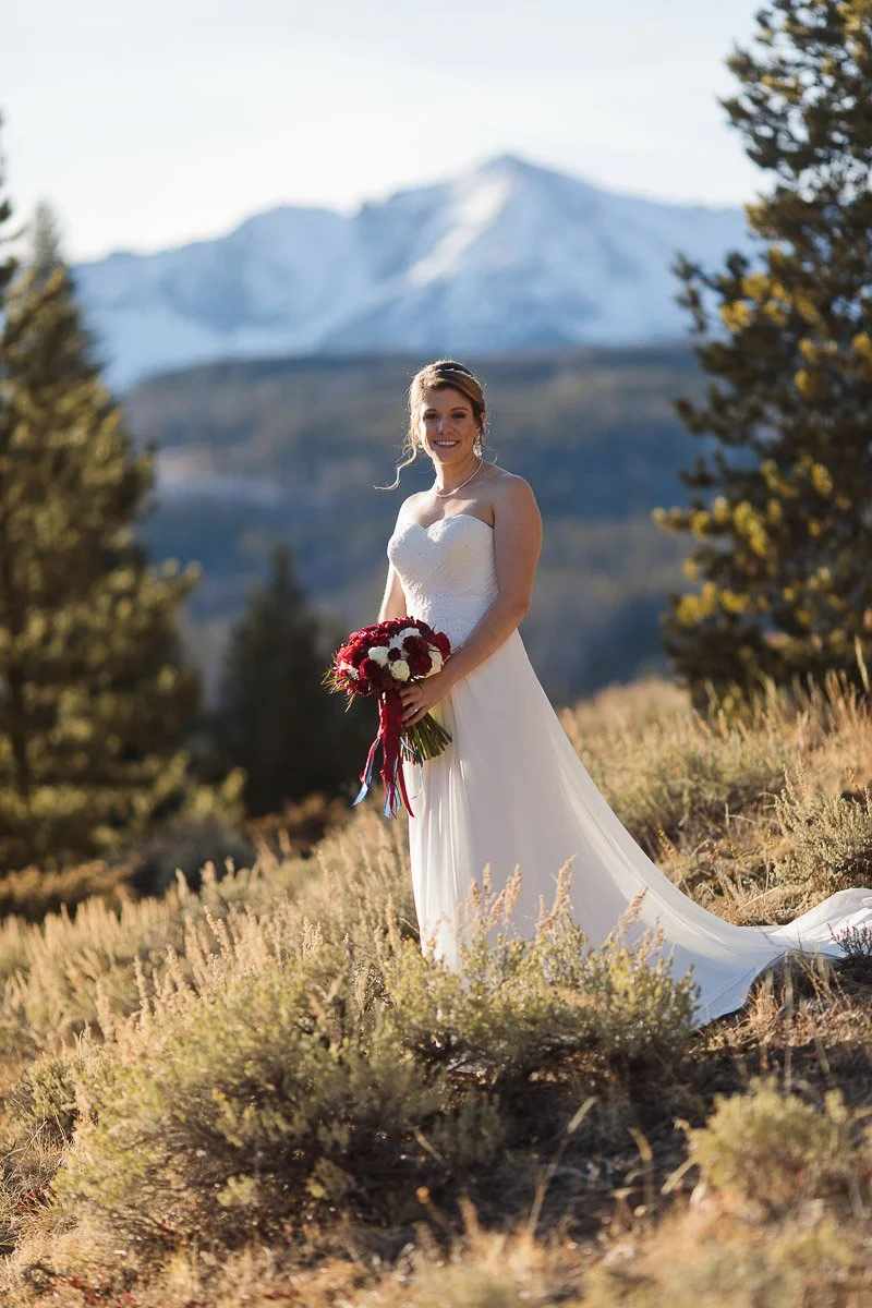 Bride in a white gown holds a red bouquet, standing in a sunlit mountain landscape. Snow-capped peaks and greenery create a serene, joyous ambiance captured by Breckenridge wedding photographer tomKphoto