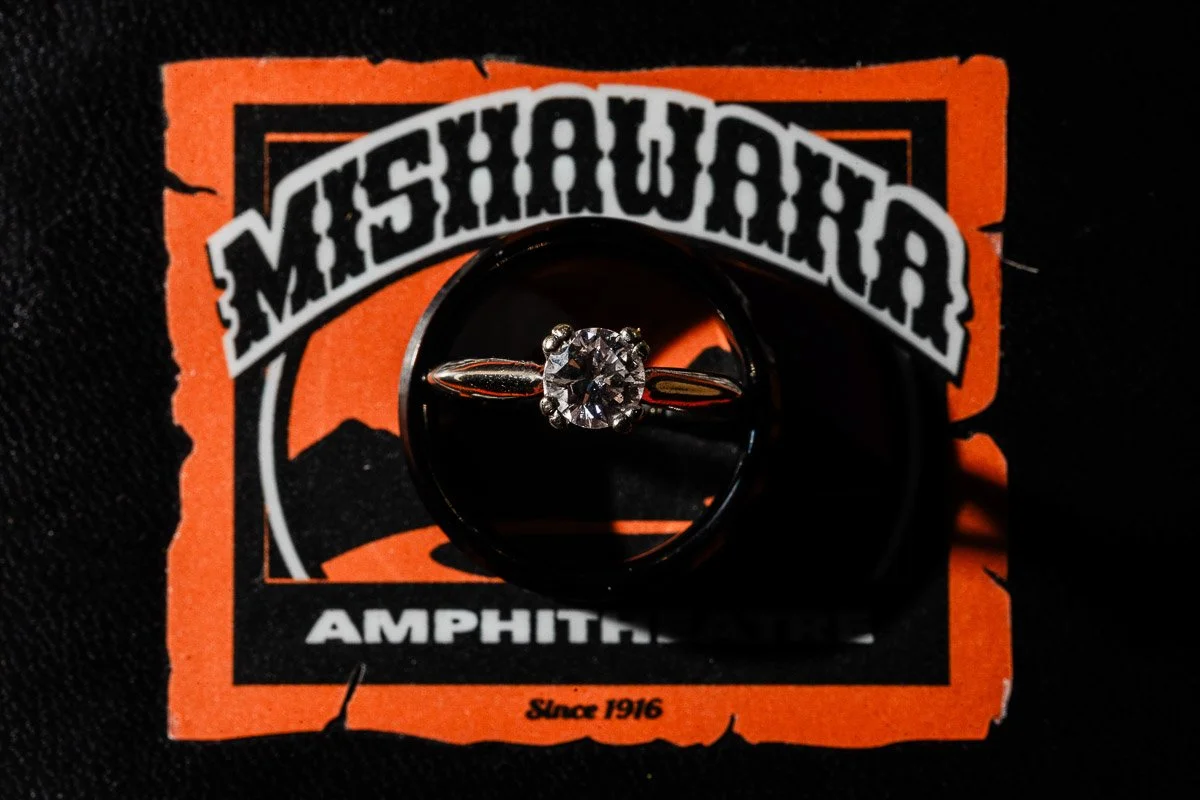 A diamond ring sits atop a black surface with a bold orange and white Mishawaka Amphitheatre logo, featuring text, curves, and shadow contrast.