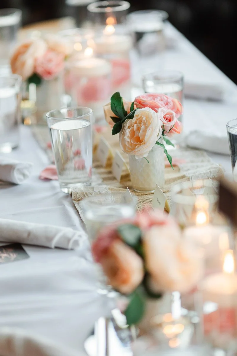 Elegant table setting with soft candlelight and pink floral arrangements in small vases. Glasses of water and neatly folded napkins add a refined touch.