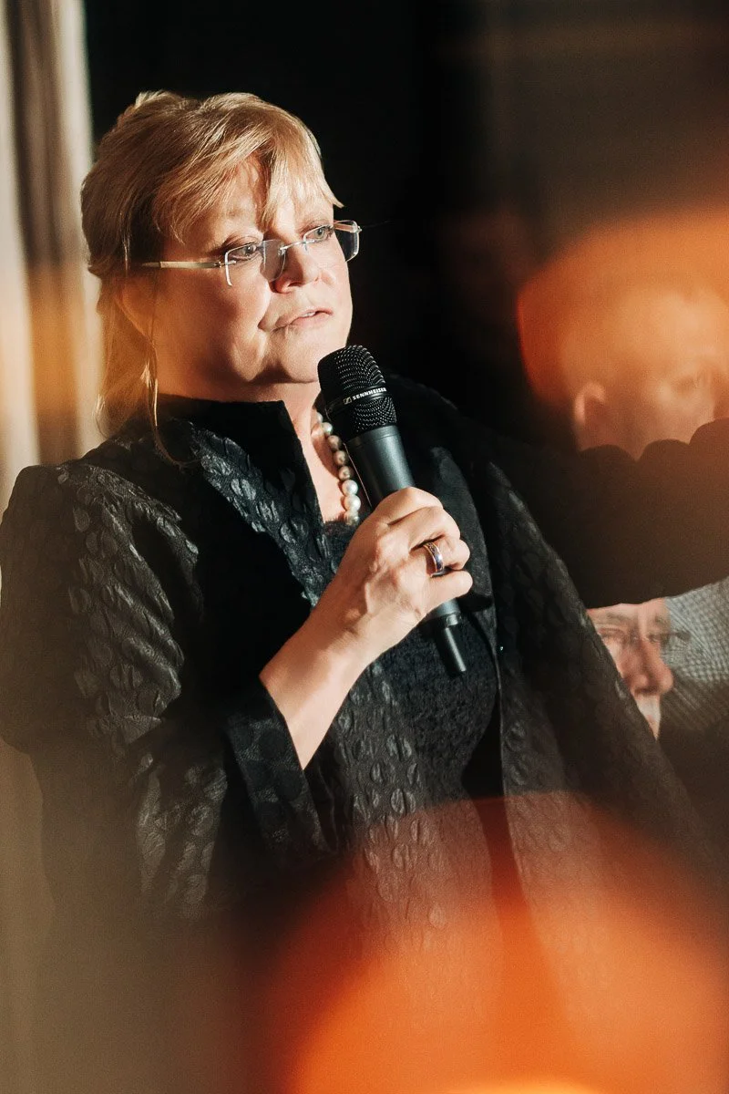 A woman with glasses, holding a microphone, speaks confidently at an event. She wears a dark outfit and pearl necklace, illuminated by warm lighting.