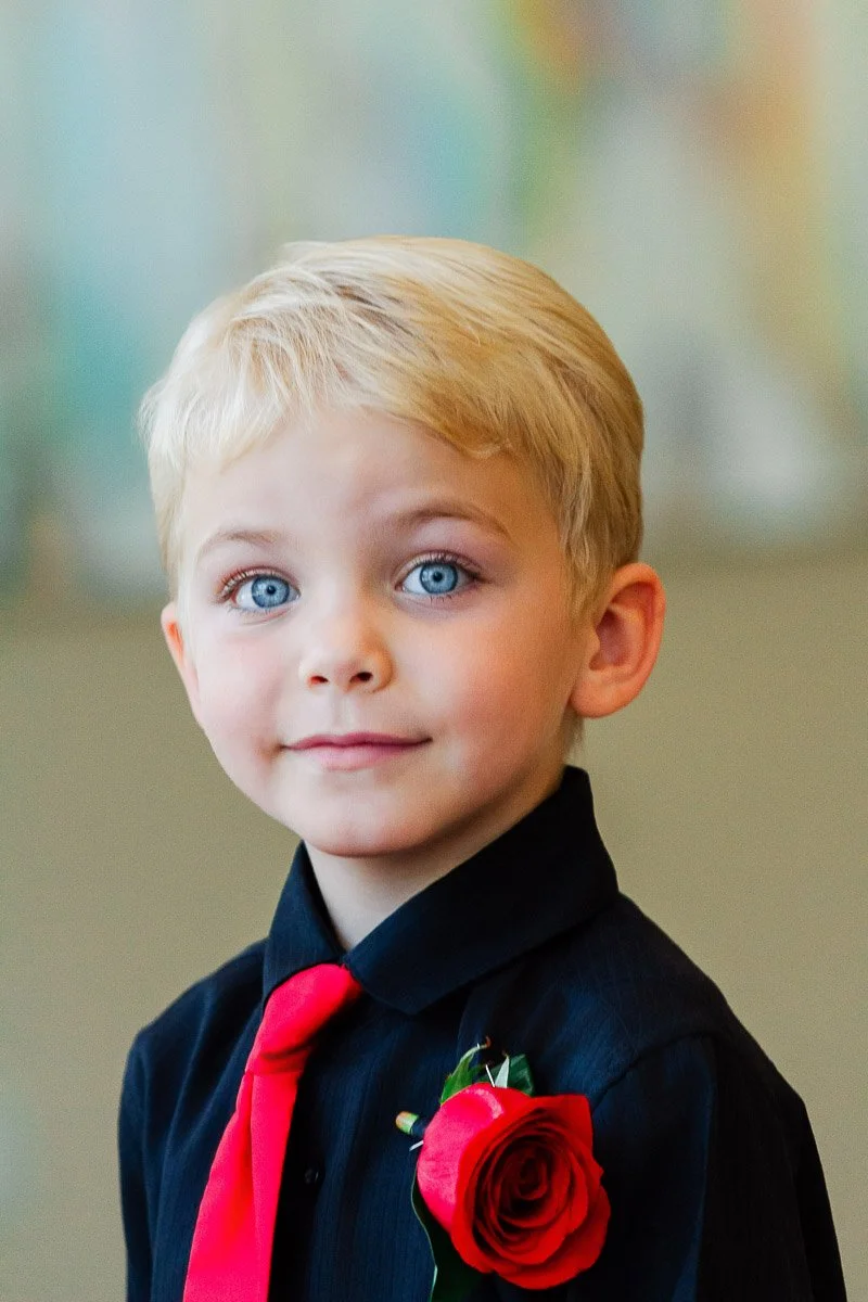 Young boy with blond hair and bright blue eyes, wearing a black shirt and red tie. A red rose is pinned to his shirt, with a soft, colorful background.