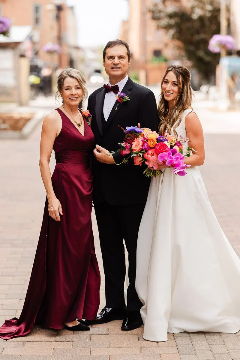 A bride in a white gown, holding vibrant pink and purple flowers, stands smiling with a man in a tuxedo and a woman in a maroon dress, outdoors.