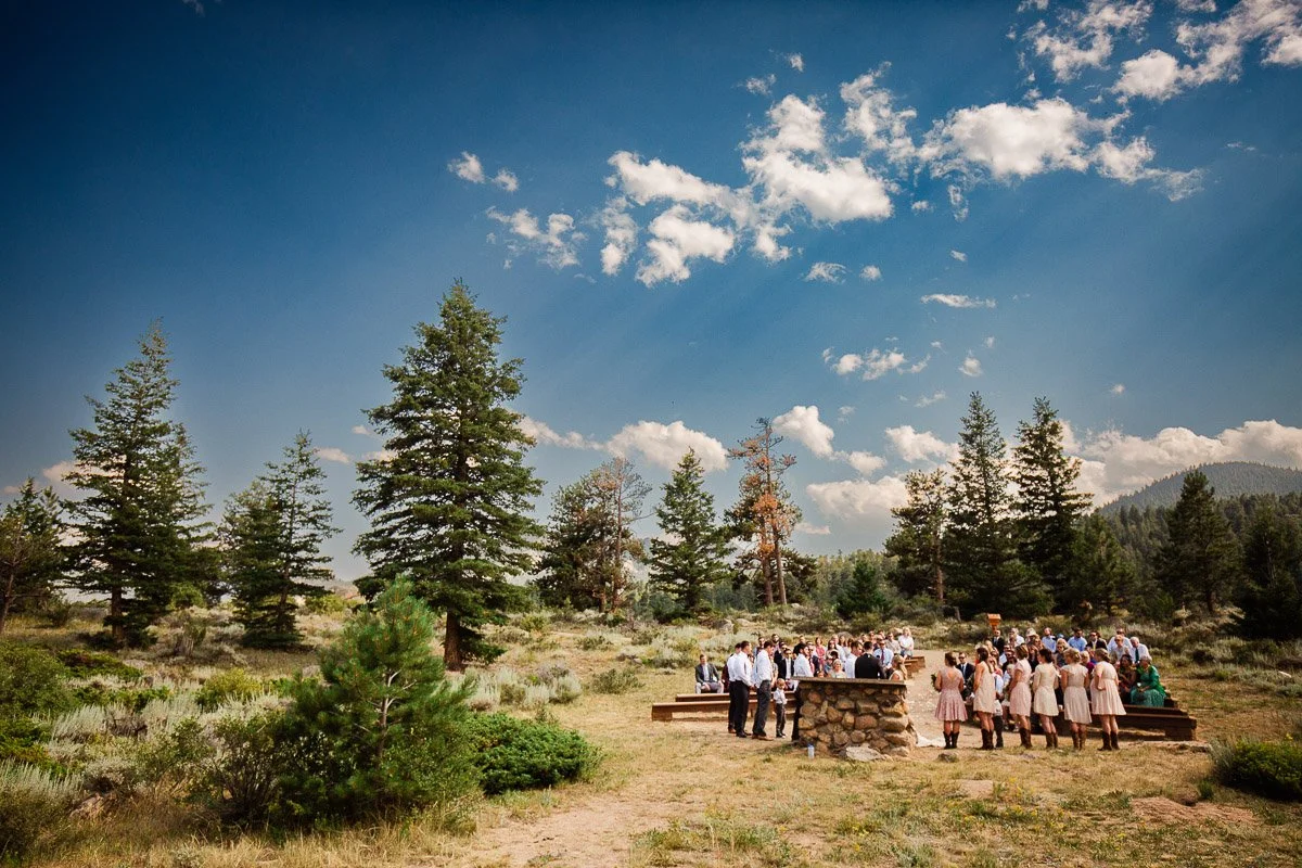 A wedding ceremony in a lush meadow at YMCA of the Rockies. Guests in formal attire gather near a rustic stone altar. Tall pine trees and a blue sky with clouds surround the scene.