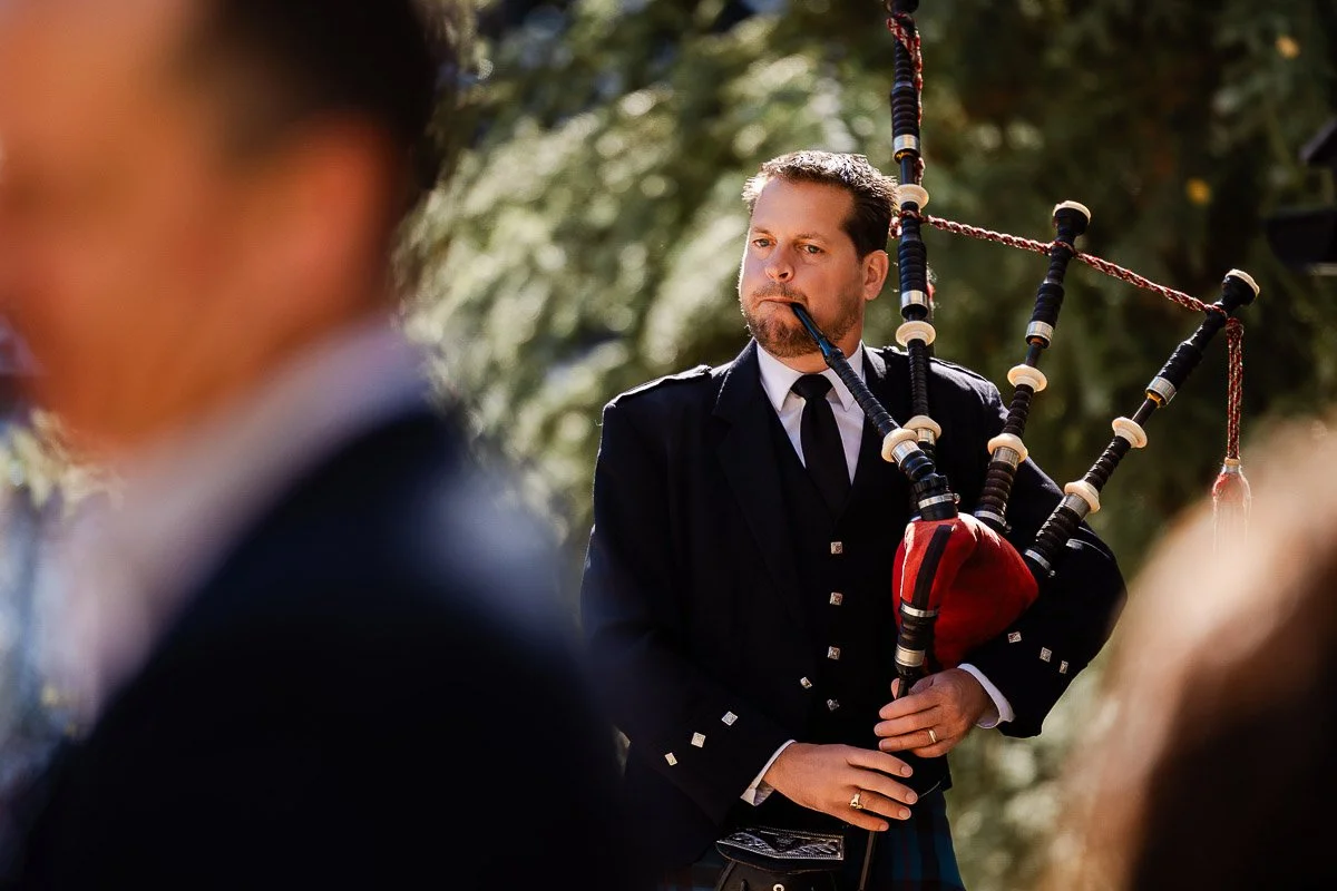 A man in traditional attire plays bagpipes outdoors, with a blurred figure in the foreground. The sunlight filters through trees, creating a serene, focused atmosphere.