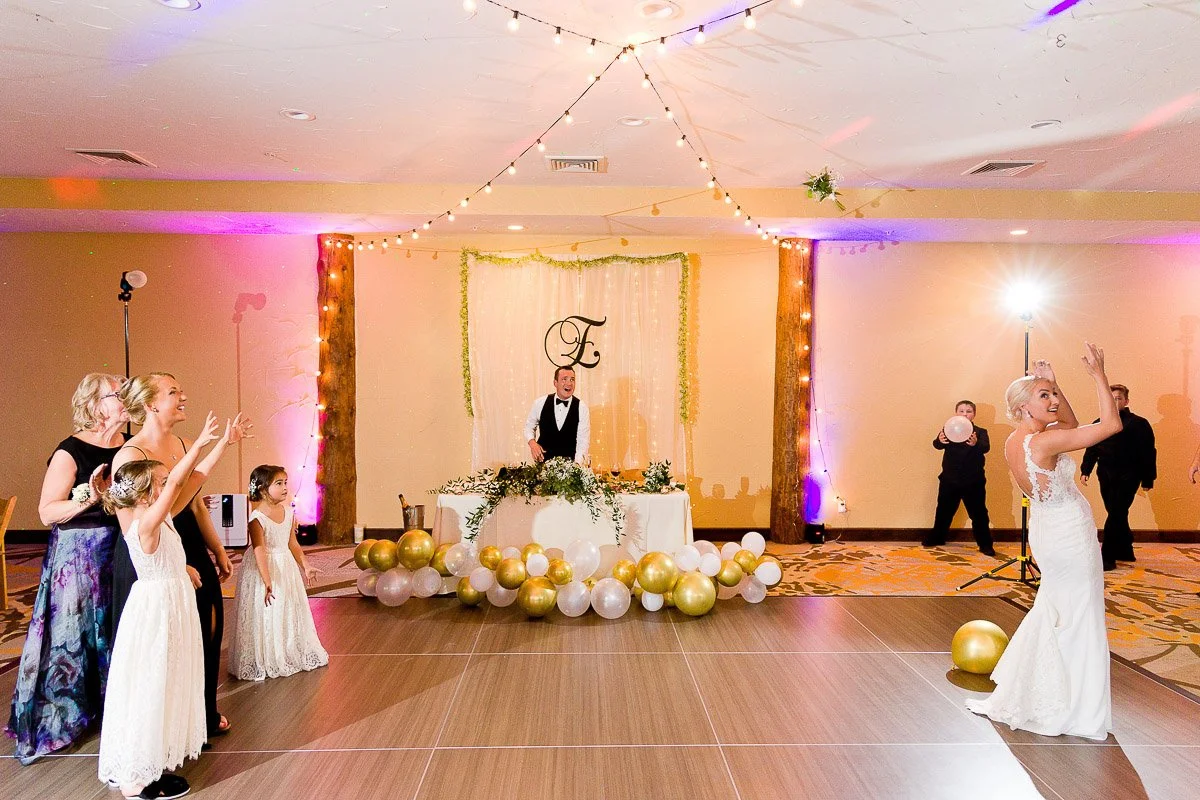 A bride in a white dress tosses her bouquet on a dance floor. Guests, including children in white dresses, excitedly reach out. Gold and white balloons adorn the room.