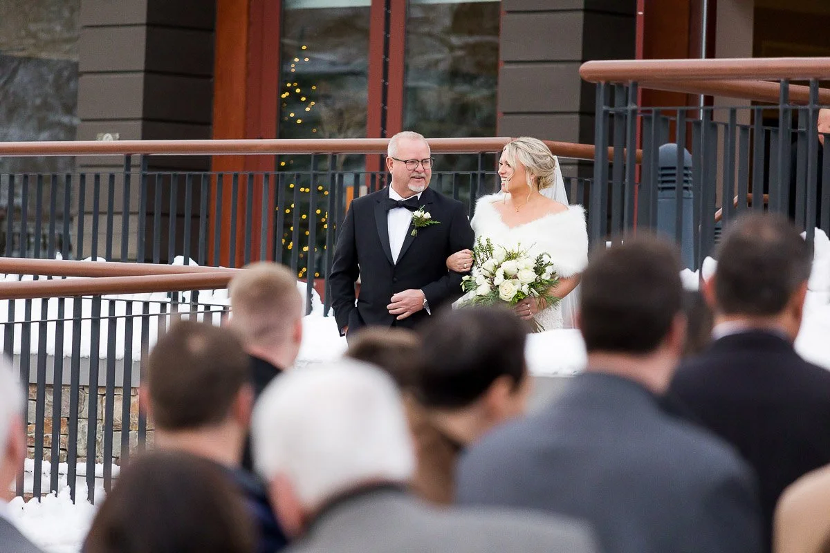 A bride and a man in a tuxedo walk arm-in-arm down an outdoor aisle. The bride holds white flowers, and snow surrounds the scene, creating a joyful, wintery atmosphere.