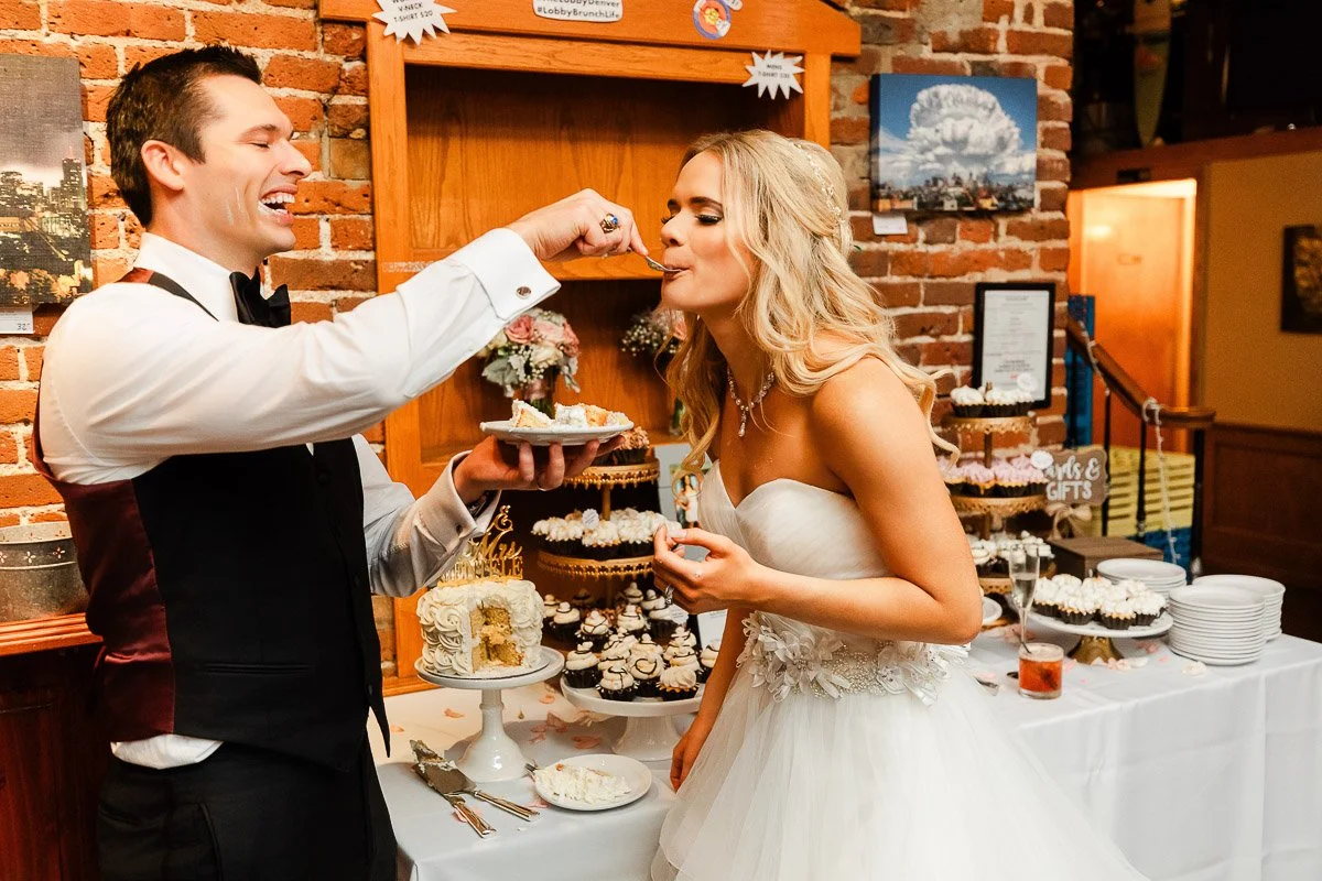 A smiling groom in a tuxedo feeds cake to a joyful bride in a white gown. They stand by a table adorned with cakes and desserts, set against a brick wall.