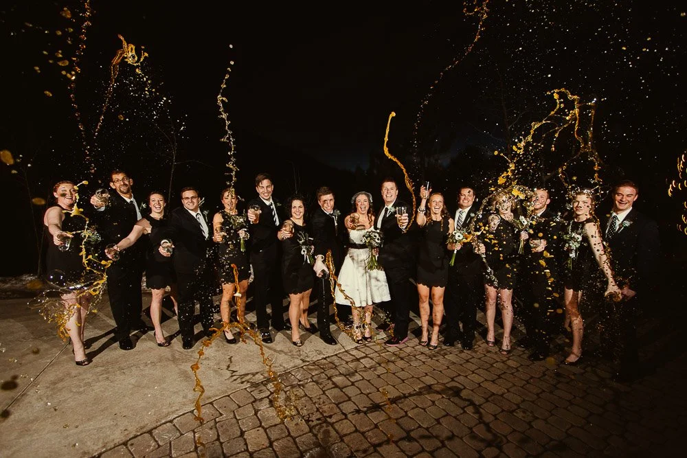 A joyful wedding party, dressed in formal black attire, celebrates outside at night. They are cheerfully spraying drinks, creating a festive, lively scene captured by Colorado Wedding Photographer tomKphoto