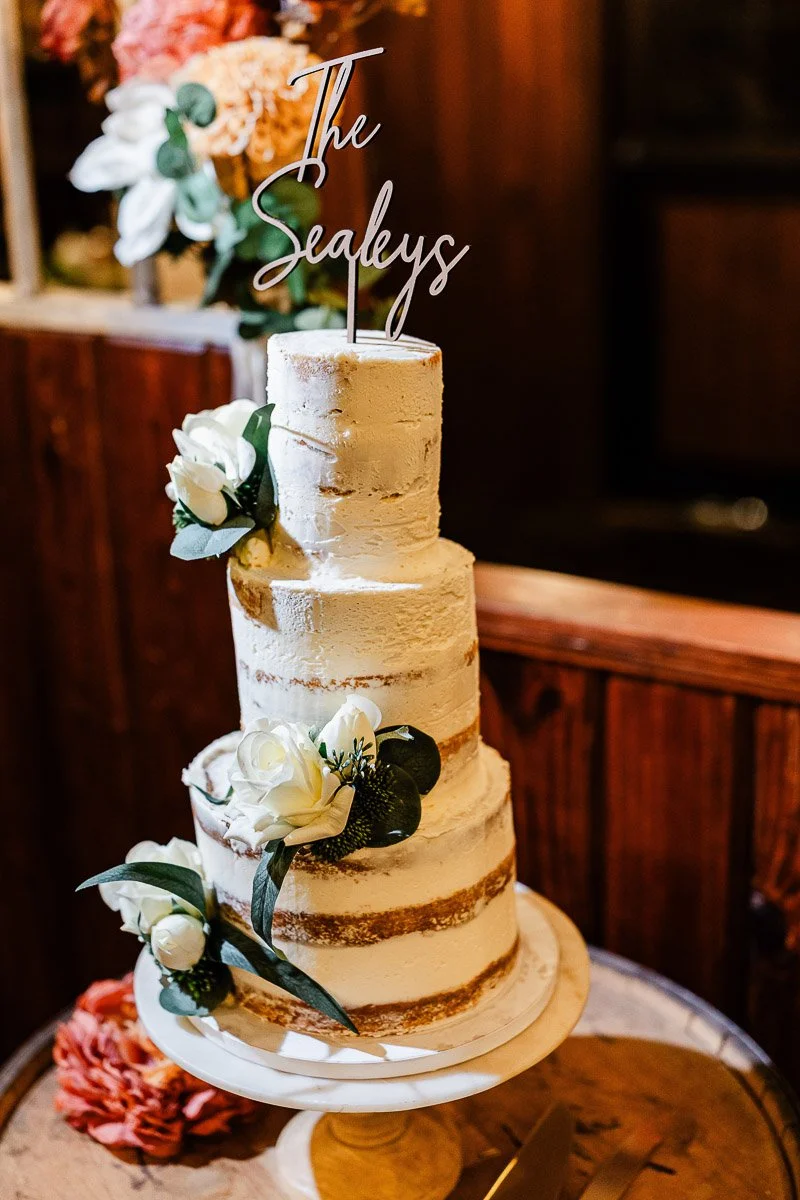 Three-tier white naked cake with white flowers and green leaves as decoration. Topped with a personalized script topper, on a wooden table.