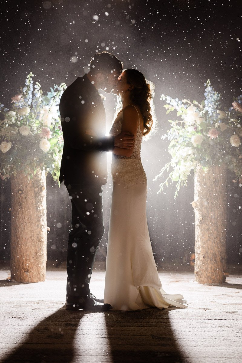 A couple stands closely in a snowy, romantic embrace, illuminated by soft backlighting. Lush floral arrangements are visible in the background, enhancing the dreamy atmosphere.