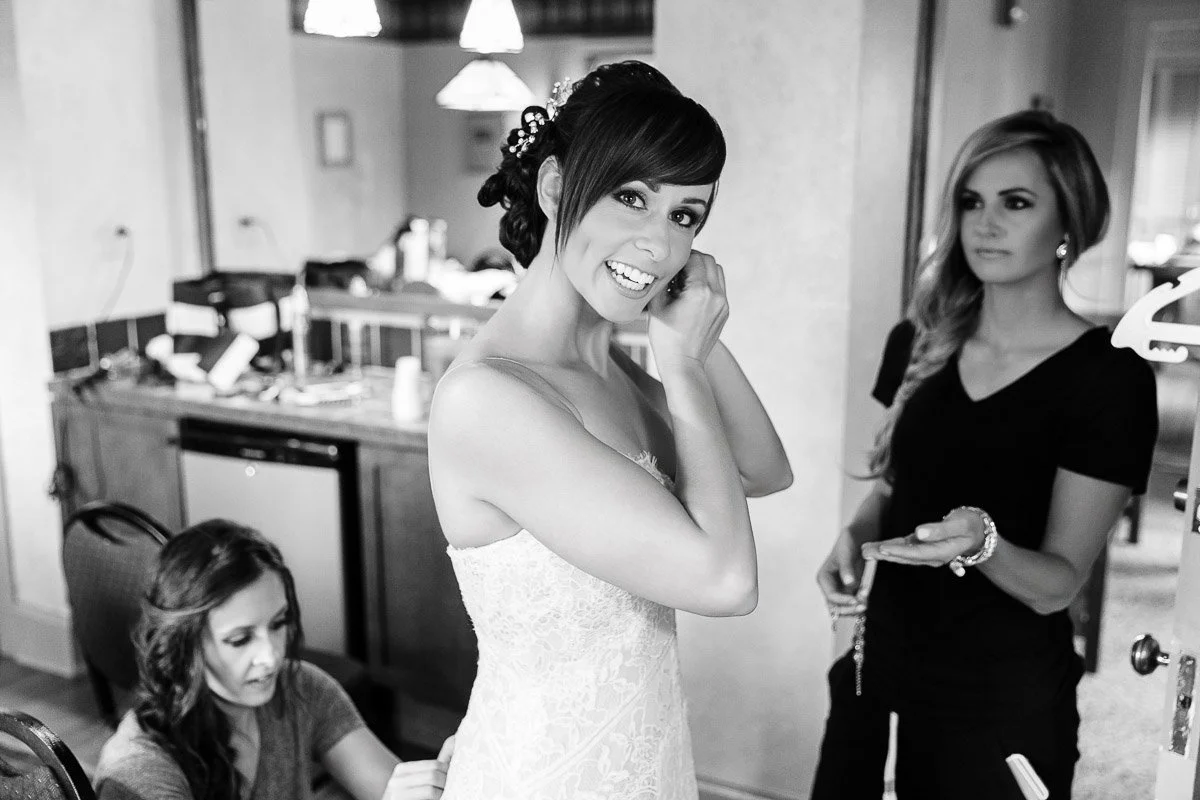 Bride smiling while adjusting her earring, wearing a lace dress. Two women assist with preparations in a warmly lit room, creating a joyful atmosphere.