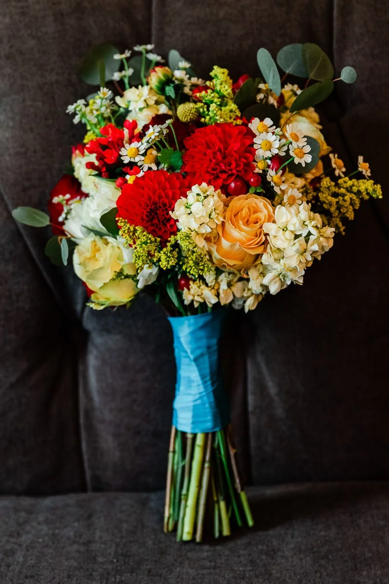 A vibrant bridal bouquet featuring red dahlias, orange roses, white hydrangeas, and daisies, wrapped in blue ribbon, on a dark gray chair.