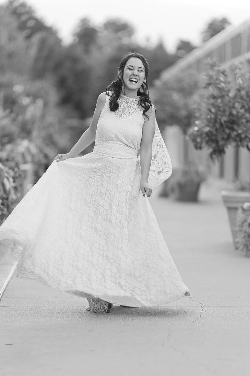 A woman joyfully twirls in a flowing lace dress, captured in black and white. She smiles brightly, exuding happiness, with blurred nature in the background.