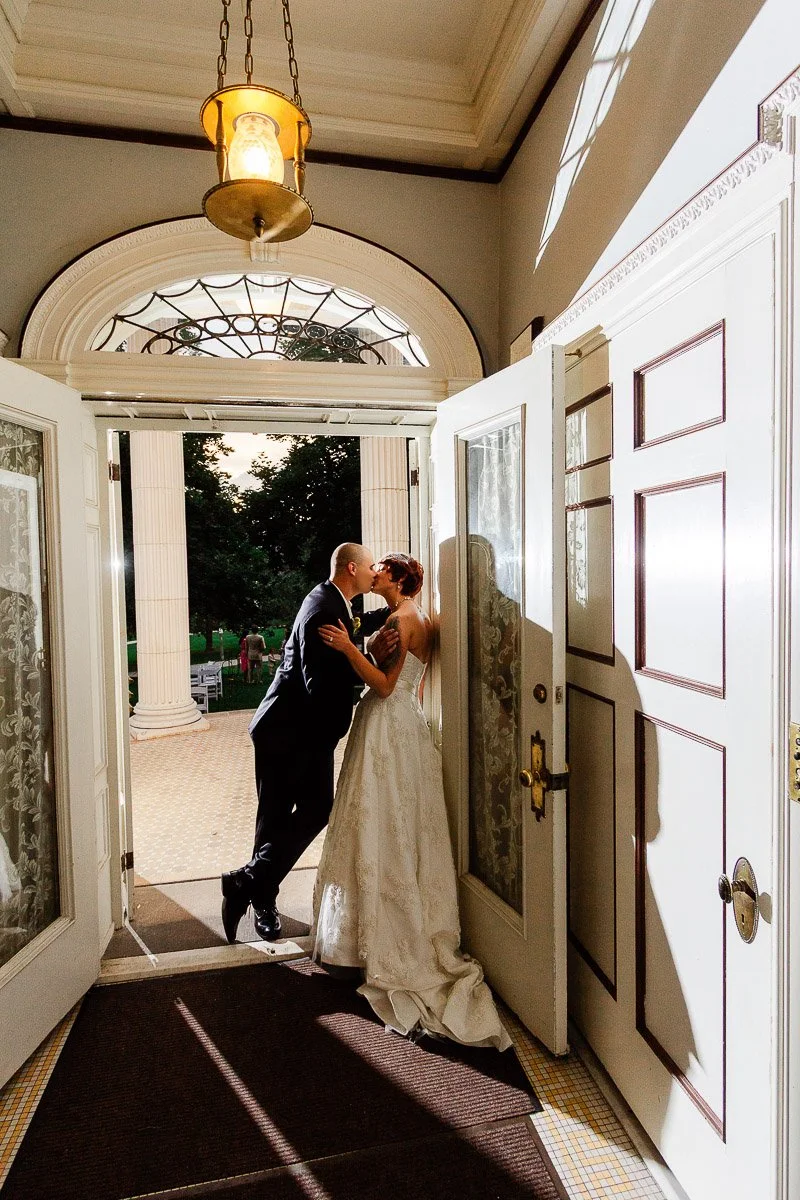 Bride and groom share a kiss at a grand entryway, framed by open doors and elegant columns. Warm light and shadows create a romantic atmosphere.