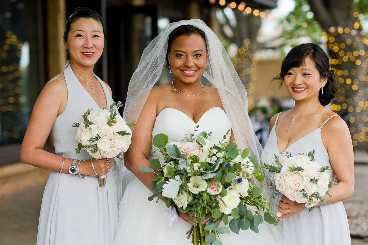 A bride smiling in a white gown and veil holds a lush bouquet, flanked by two bridesmaids in light gray dresses, each holding white flower bouquets.