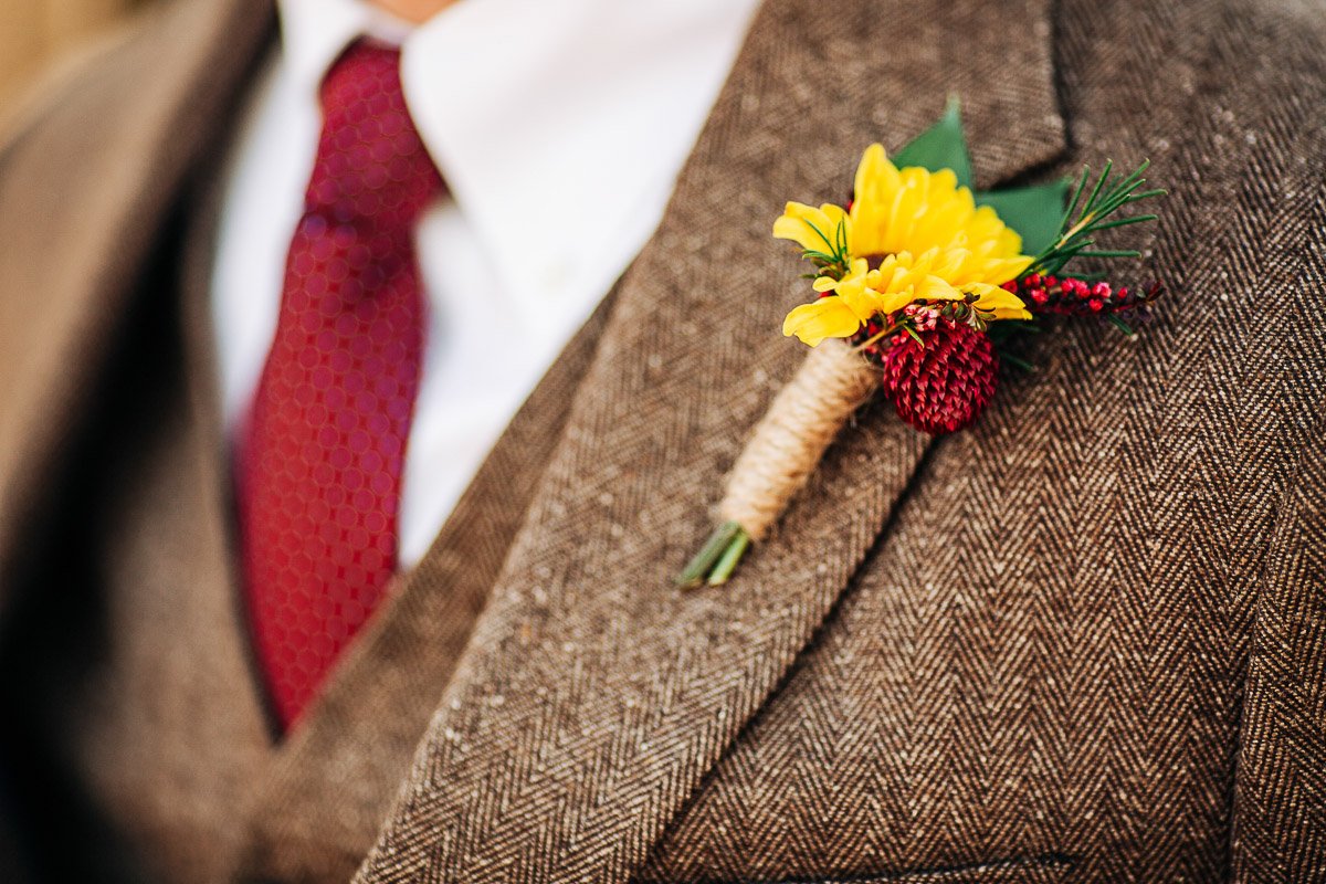 A close-up of a man in a tweed suit and red tie. The jacket features a bright boutonniere with yellow and red flowers, creating an elegant touch.