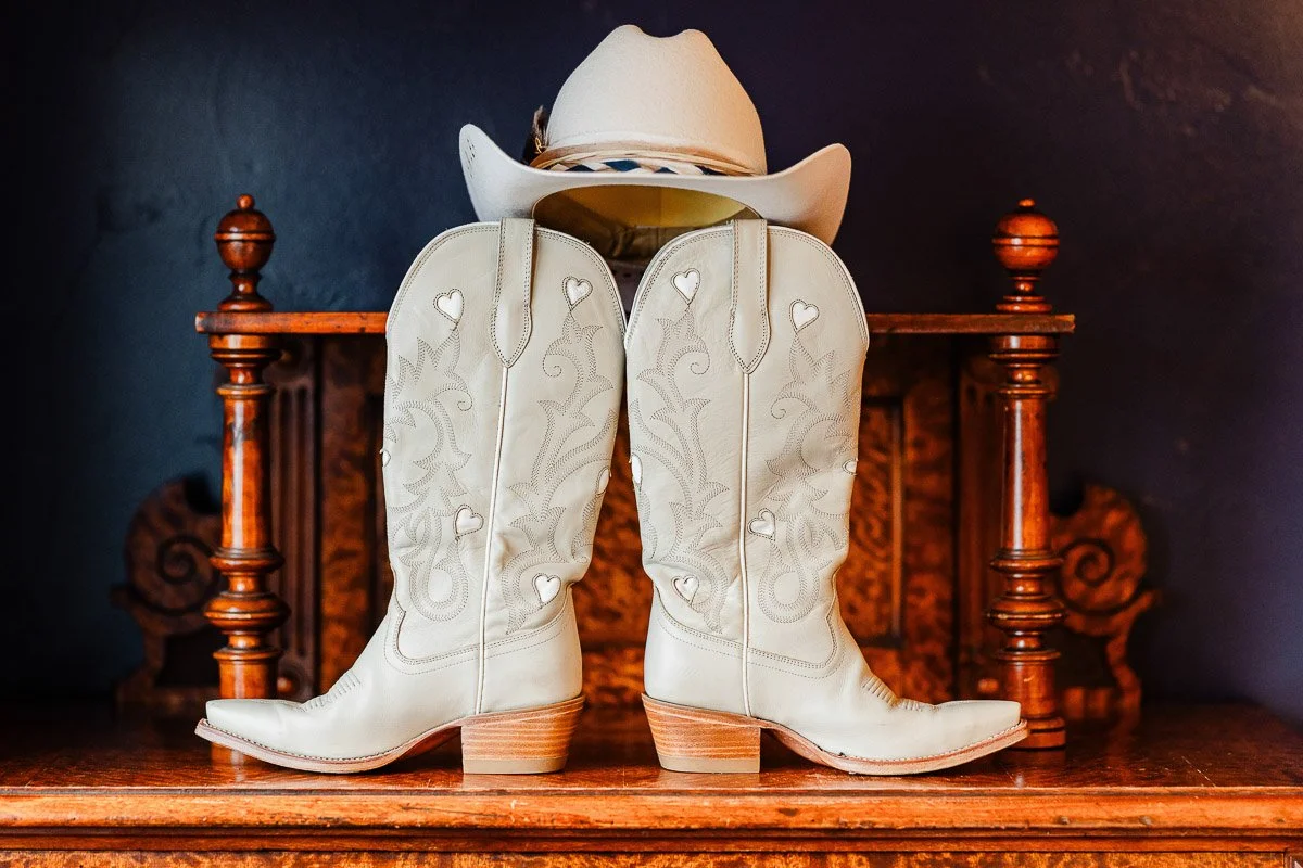 White cowboy boots with heart designs stand on a wooden dresser, topped by a matching cowboy hat. The setting conveys a rustic, country vibe.