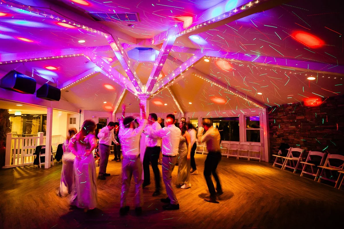 People are dancing energetically in a warmly-lit room. Colored lights and laser patterns create a festive atmosphere, reflecting joy and celebration.