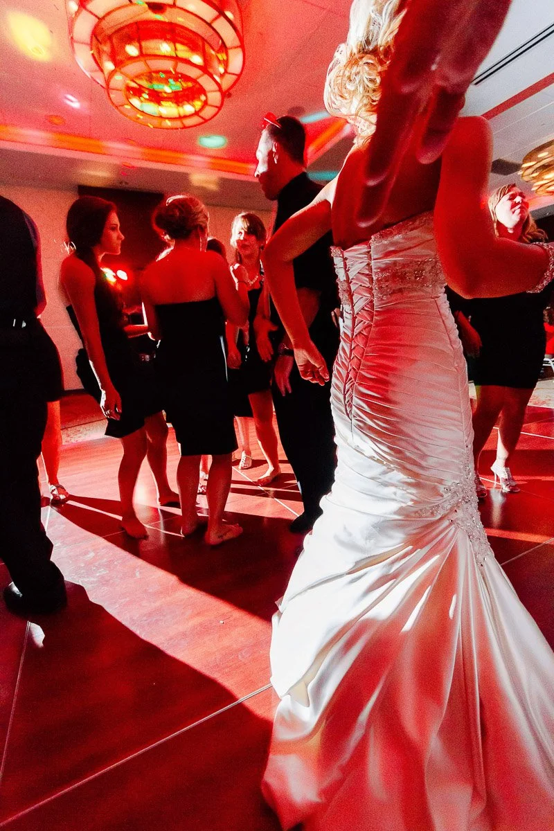 A bride in a white gown dances at a lively wedding reception. Guests in black attire surround her on a warmly lit dance floor, creating a joyful atmosphere.