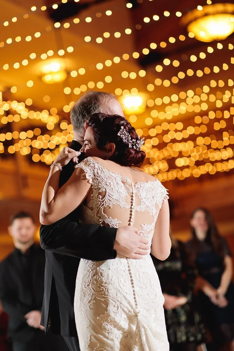 A couple embraces during a dance at a warmly lit wedding. The bride's lace gown and soft, glowing lights above create a romantic atmosphere.