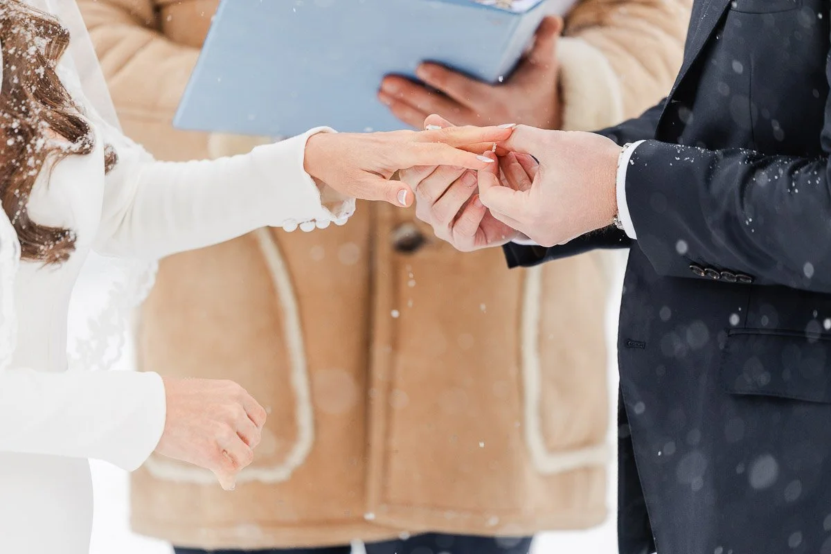 A couple exchanges rings in the snow during an outdoor wedding ceremony. The bride is in a white dress, the groom in a suit, and a person holds a folder.