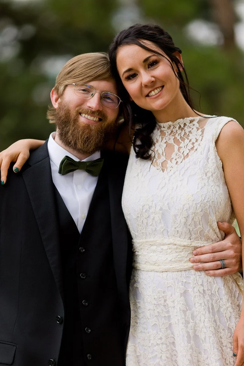A couple poses outdoors, smiling warmly. The man wears a black suit with a green bow tie, and the woman wears a white lace dress. Both appear happy and content.