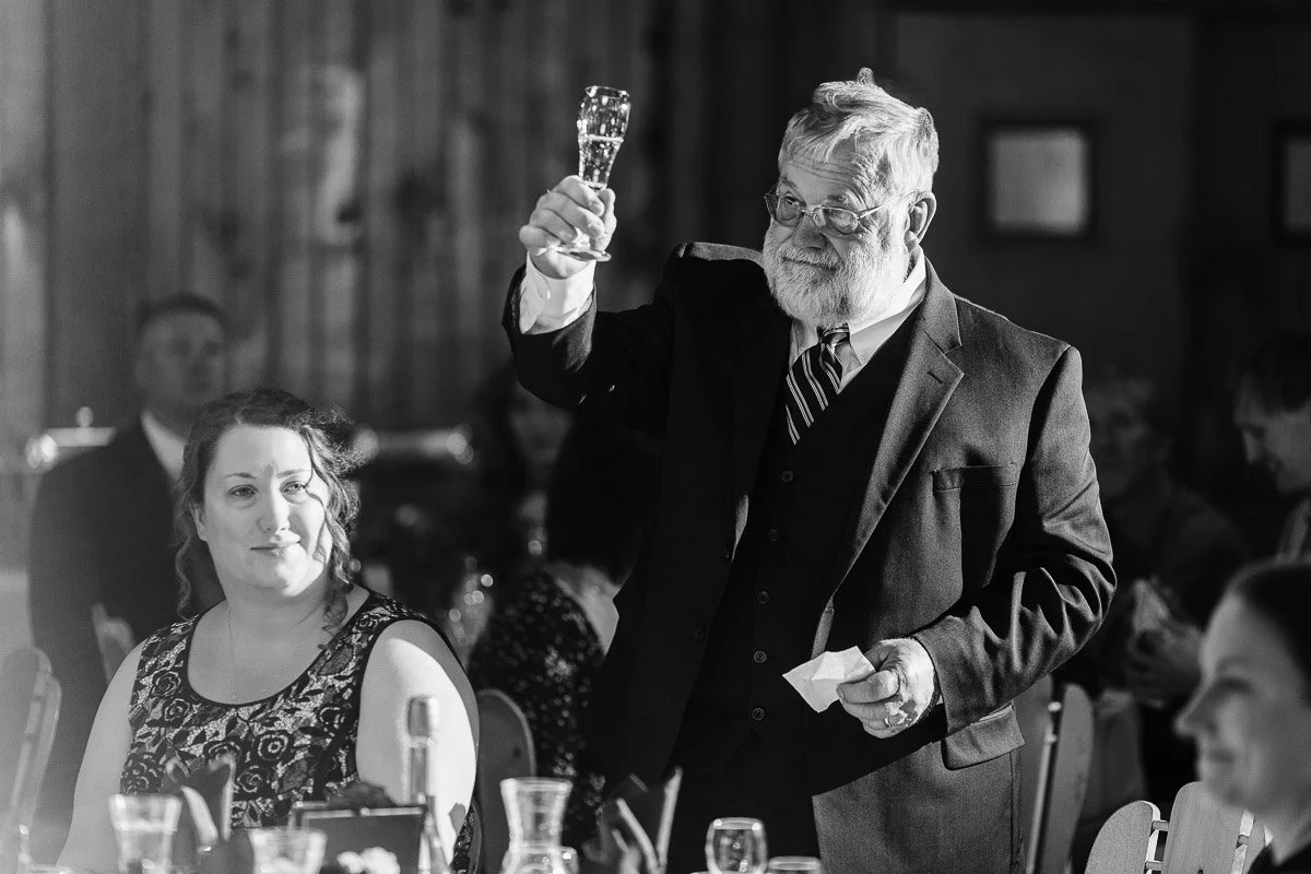 A man in a suit raises a glass for a toast at a formal event, with a warm smile. A woman sits nearby, watching. The setting is elegant and celebratory.
