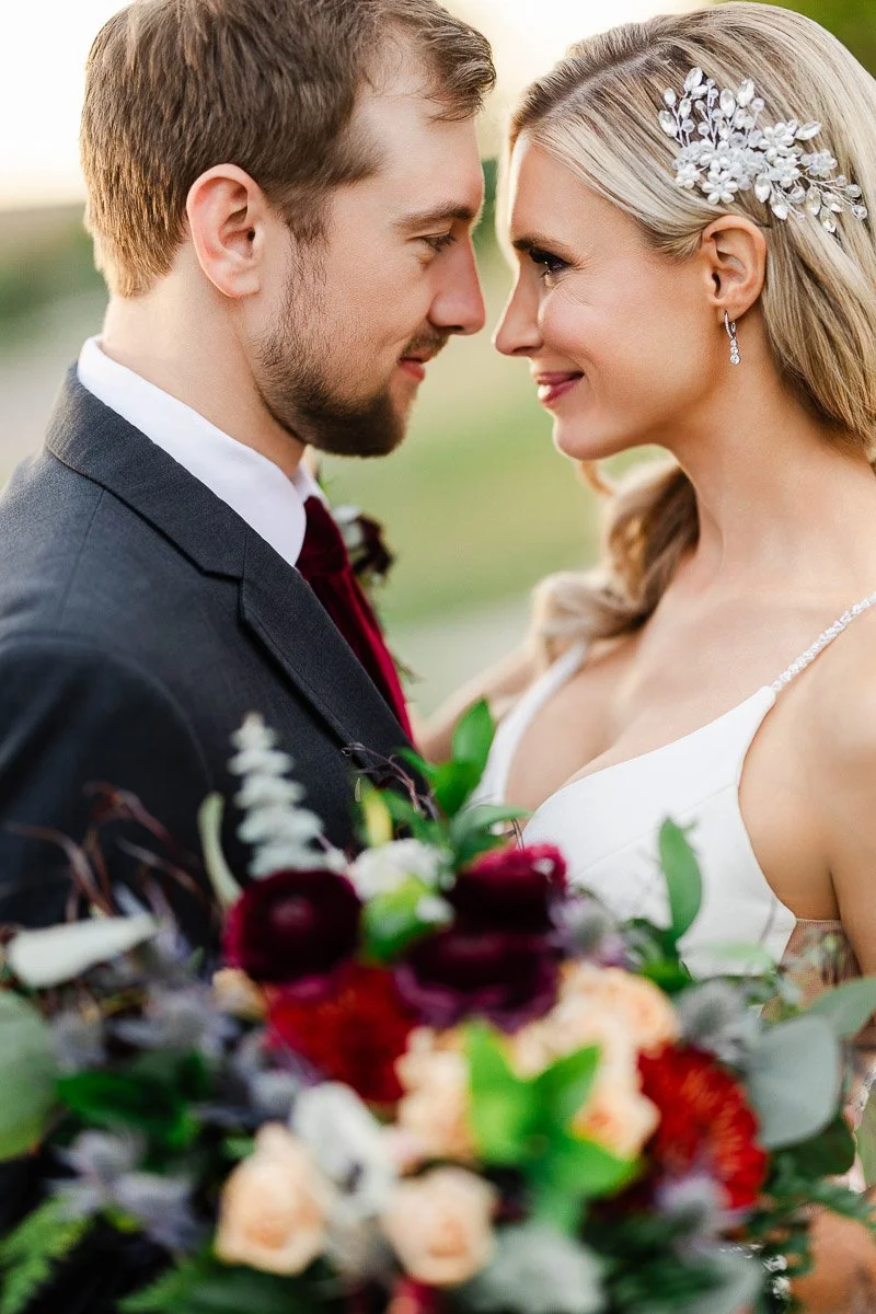 A bride and groom gaze lovingly at each other, smiling softly. She holds a colorful bouquet with red and peach flowers. The scene feels romantic and serene.