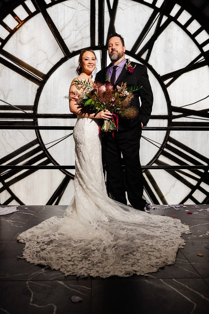 A couple in wedding attire stands smiling in front of a large clock face. The bride holds a bouquet, and the groom is in a suit. The setting feels timeless and elegant.